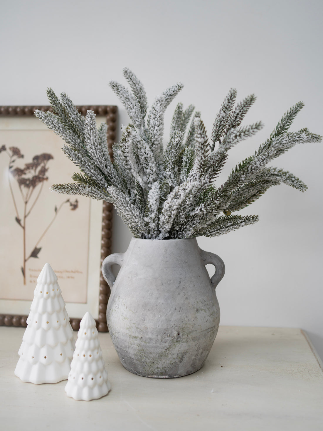 Decorative arrangement with a vase of frosted branches and two small white ceramic trees on a light surface.