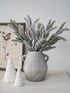 Decorative arrangement with a vase of frosted branches and two small white ceramic trees on a light surface.
