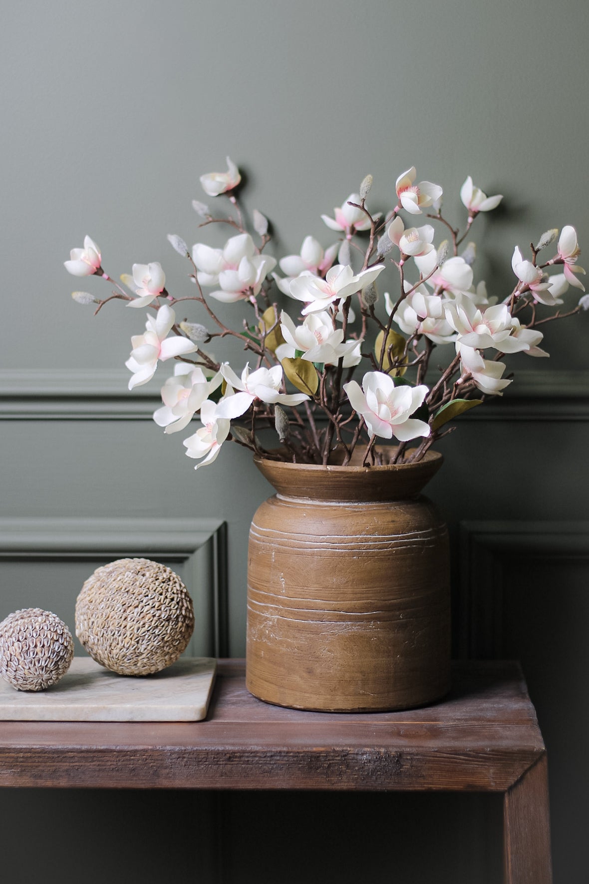 Faux Soft Pink Magnolia Stems in brown rustic vase on wooden table with decorative shell spheres