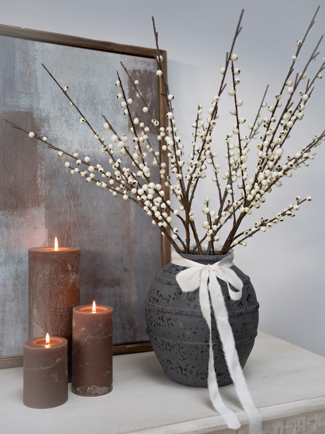 Decorative arrangement with a vase of branches, candles, and a framed picture on a surface.
