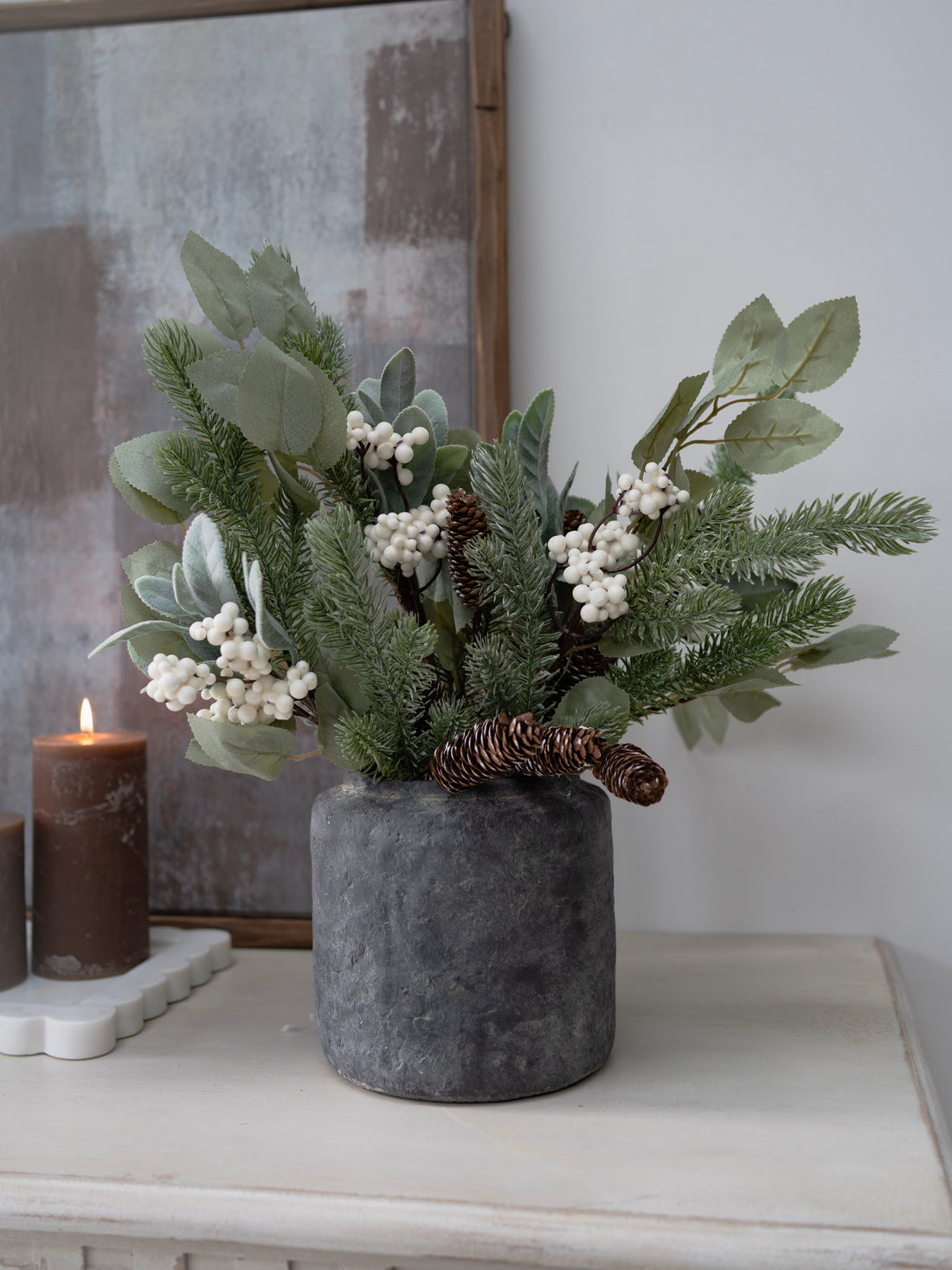 Decorative arrangement of faux christmas greenery and berries in a gray vase on a surface with a candle and framed picture in the background.