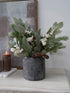 Decorative arrangement of faux christmas greenery and berries in a gray vase on a surface with a candle and framed picture in the background.