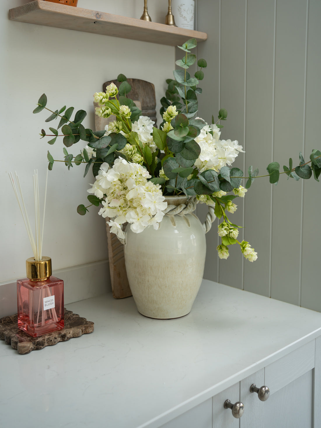White vase with Faux White Butterfly Hydrangeas, Hops and Eucalyptus Floral Arrangement on a white surface next to a pink diffuser on brown marble tray