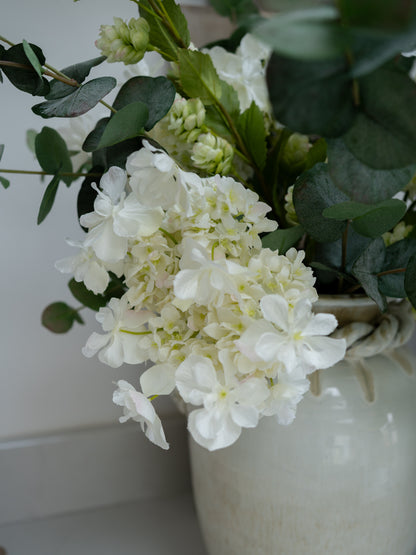 White faux hydrangeas flowers in a vase with green leaves on a white background