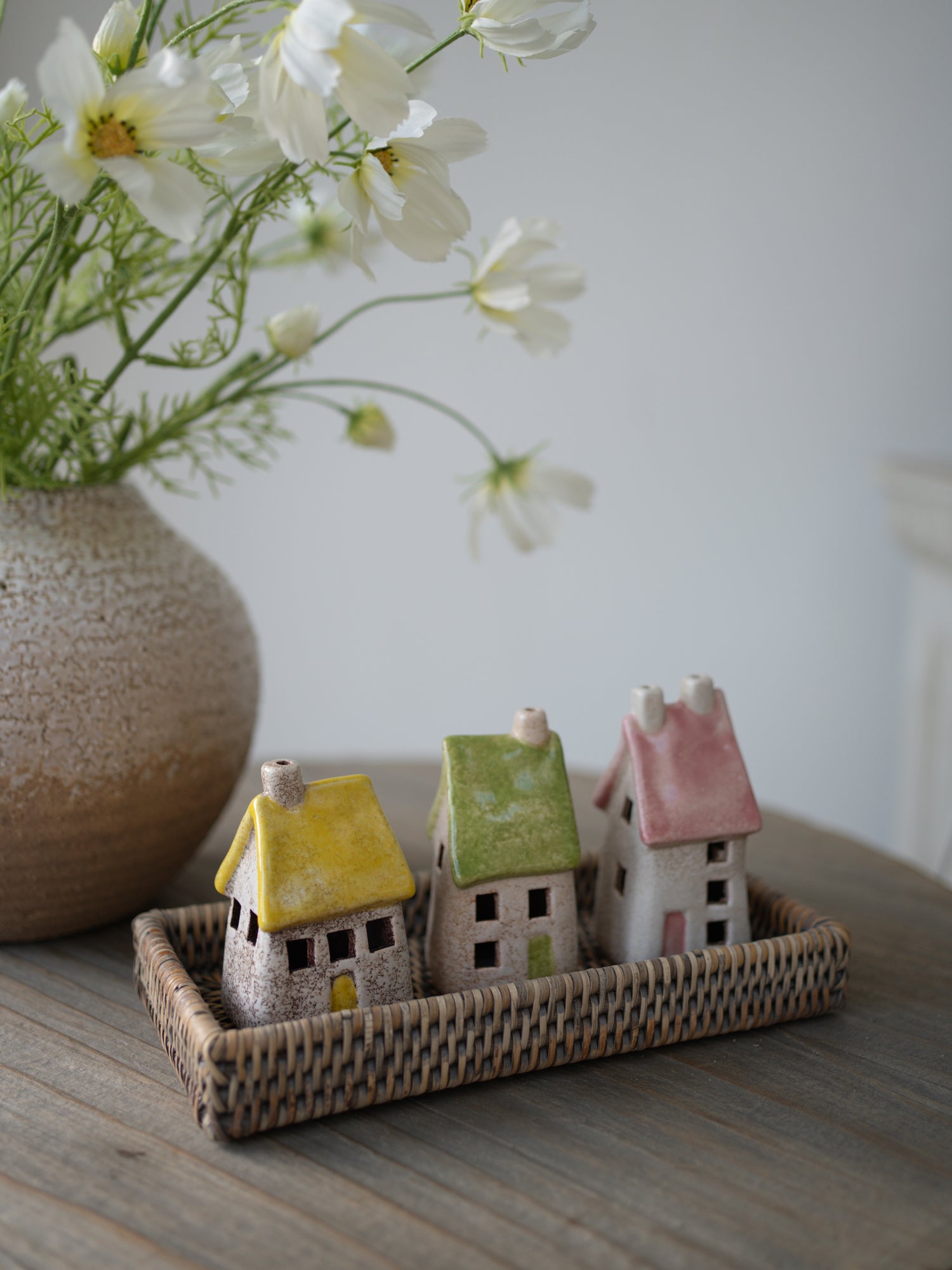 pastel toned roof stoneware houses on grey washed rattan tray with white faux cosmos and vase in the background on grey wooden table