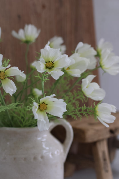 faux White cosmos flowers in a white vase on a wooden surface