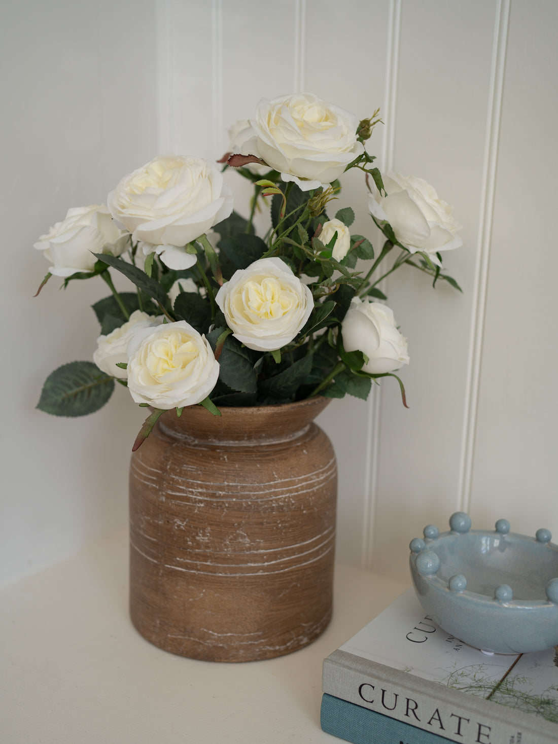 Bouquet of white roses in a rustic brown vase on a shelf with books.