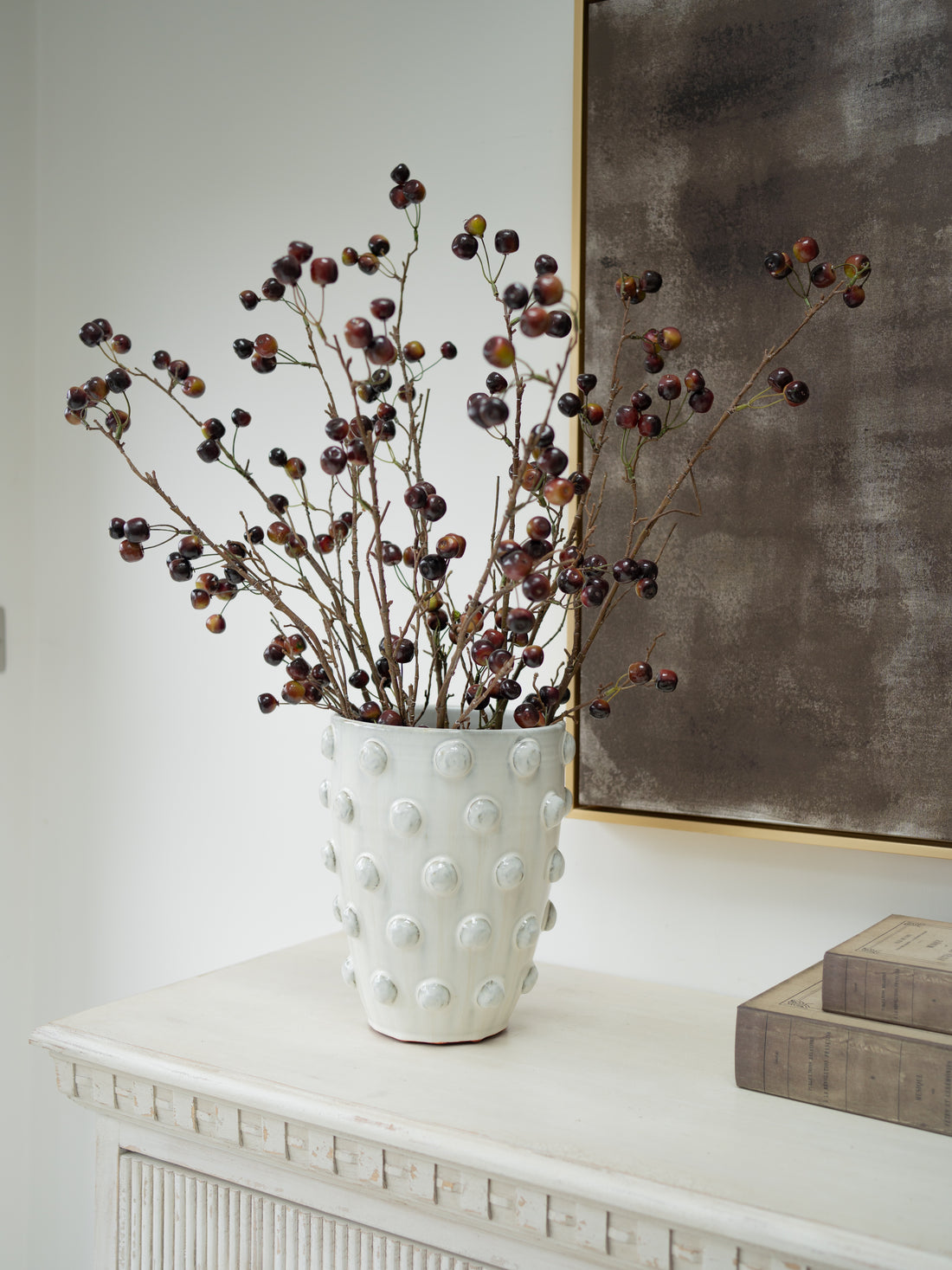 A white textured ceramic vase with a raised bubble pattern, filled with faux wild cherry branches, styled on a white console table next to stacked books and a framed artwork.
