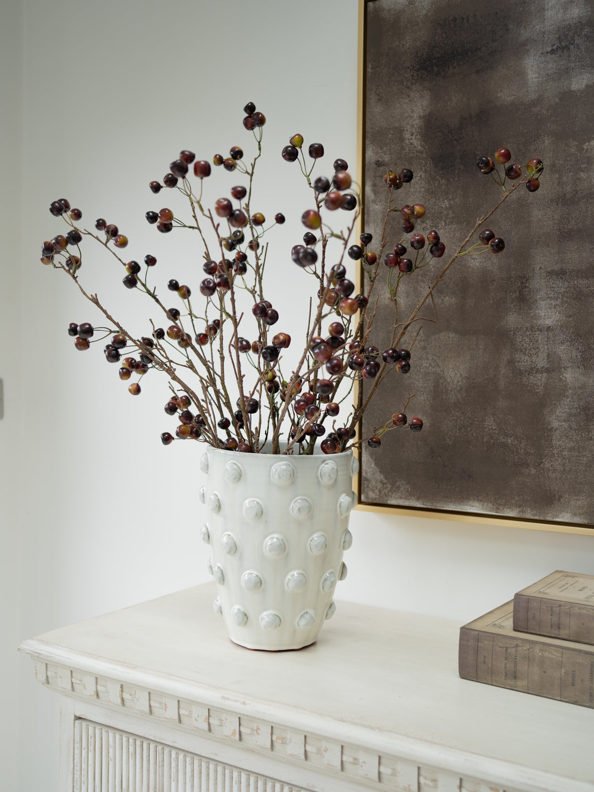 A white textured ceramic vase with a raised bubble pattern, filled with faux wild cherry branches, styled on a white console table next to stacked books and a framed artwork.