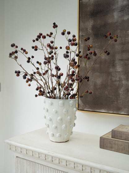 A white textured ceramic vase with a raised bubble pattern, filled with faux wild cherry branches, styled on a white console table next to stacked books and a framed artwork.
