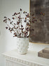A white textured ceramic vase with a raised bubble pattern, filled with faux wild cherry branches, styled on a white console table next to stacked books and a framed artwork.