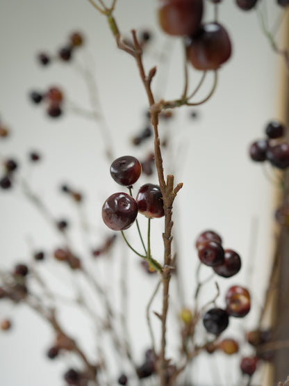 Faux autumn arrangement featuring dark brown and black cherry-like berries on slender brown branches, photographed against a soft off-white background.