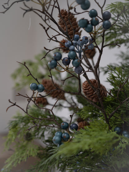Natural christmas greenery arrangement featuring green cedar-like branches with clusters of blue-gray berries and brown pine cones against a soft neutral background.