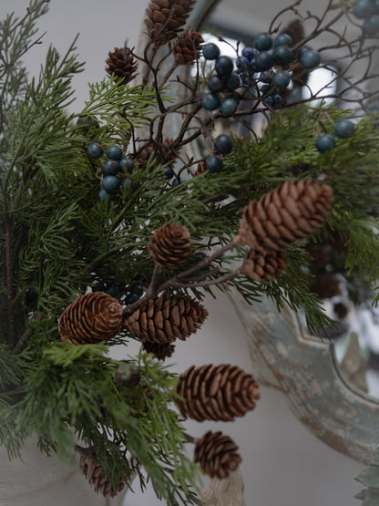 Faux pinecones and blue berries garland with green evergreen branches, shown in a close-up decorative shot.
