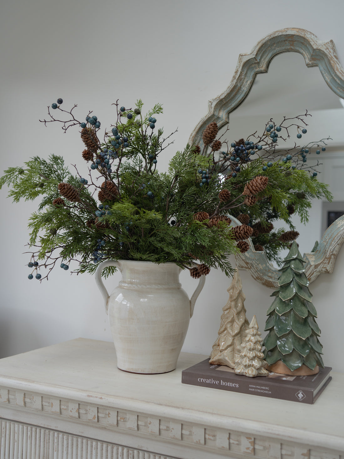 A rustic ceramic pitcher vase filled with a winter evergreen arrangement of pine branches, pinecones, and blue berries, styled on a white console table next to decorative tree figurines and an ornate mirror.