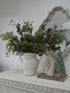 A rustic ceramic pitcher vase filled with a winter evergreen arrangement of pine branches, pinecones, and blue berries, styled on a white console table next to decorative tree figurines and an ornate mirror.