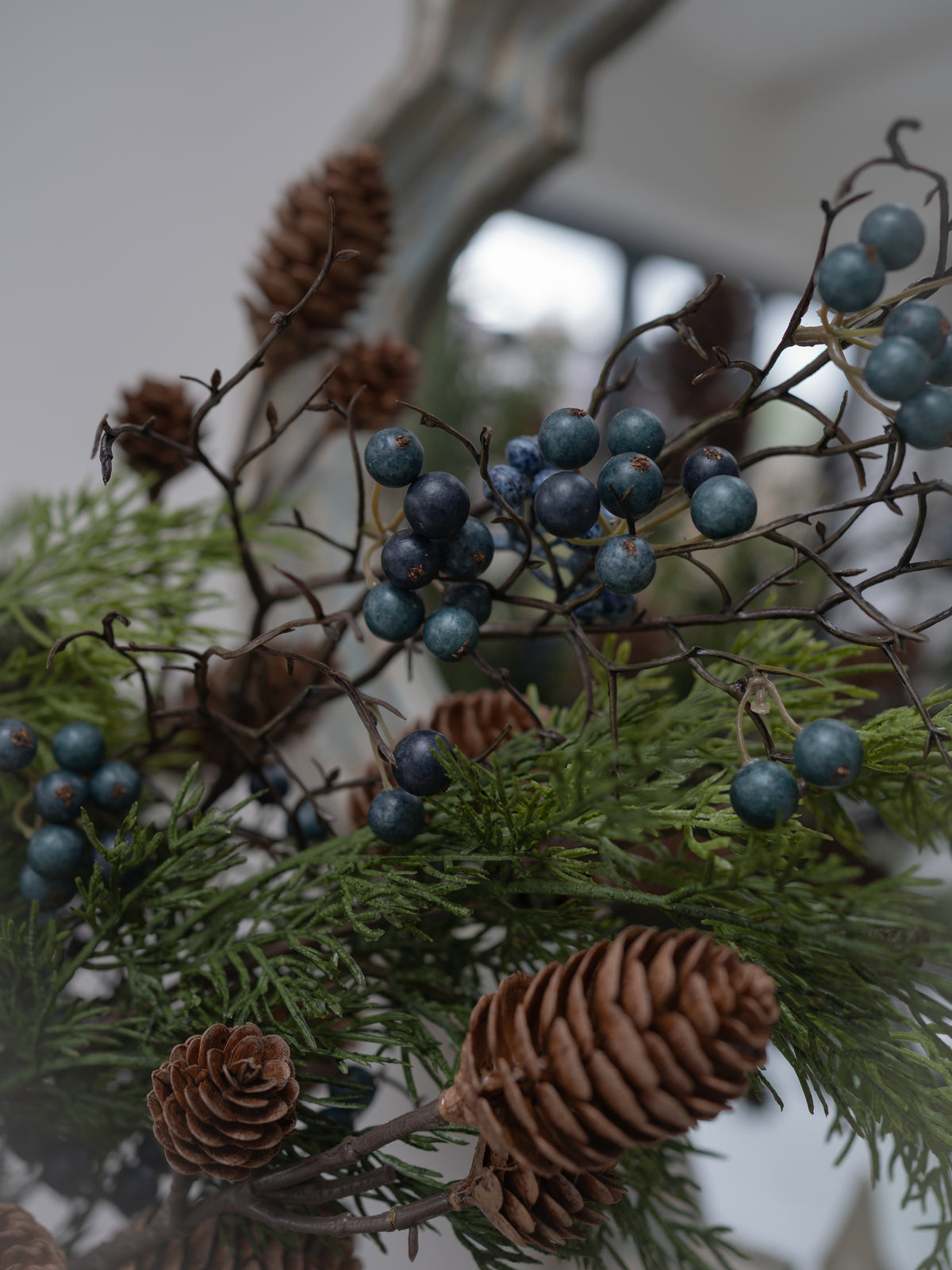 Natural Christmas greenery arrangement featuring pinecones, evergreen branches, and clusters of dark blue berries on thin brown branches.