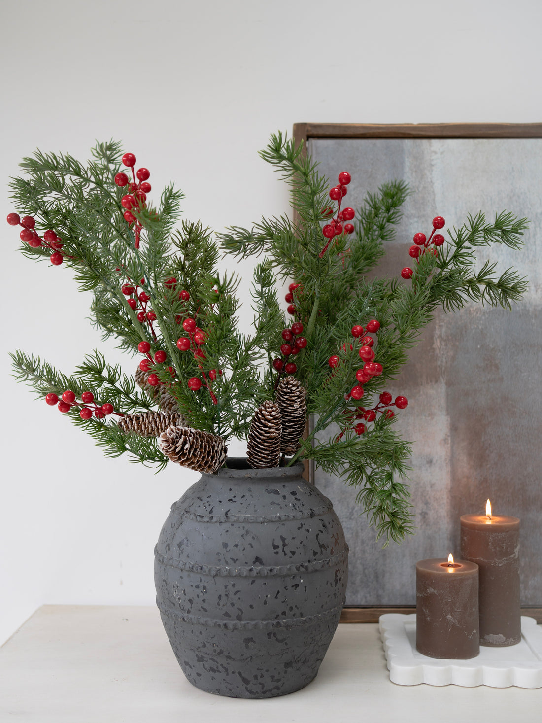 Decorative arrangement with faux christmas green branches and red berries in a textured gray vase on a white surface.