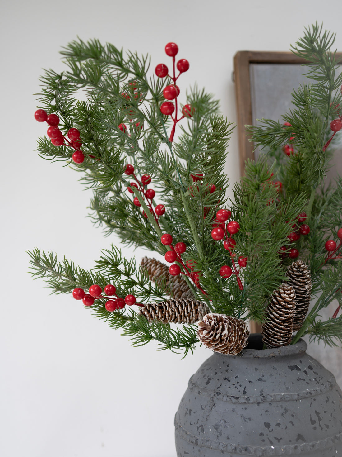 Decorative arrangement with faux christmas greenery, red berries, and pinecones in a gray vase against a white wall.