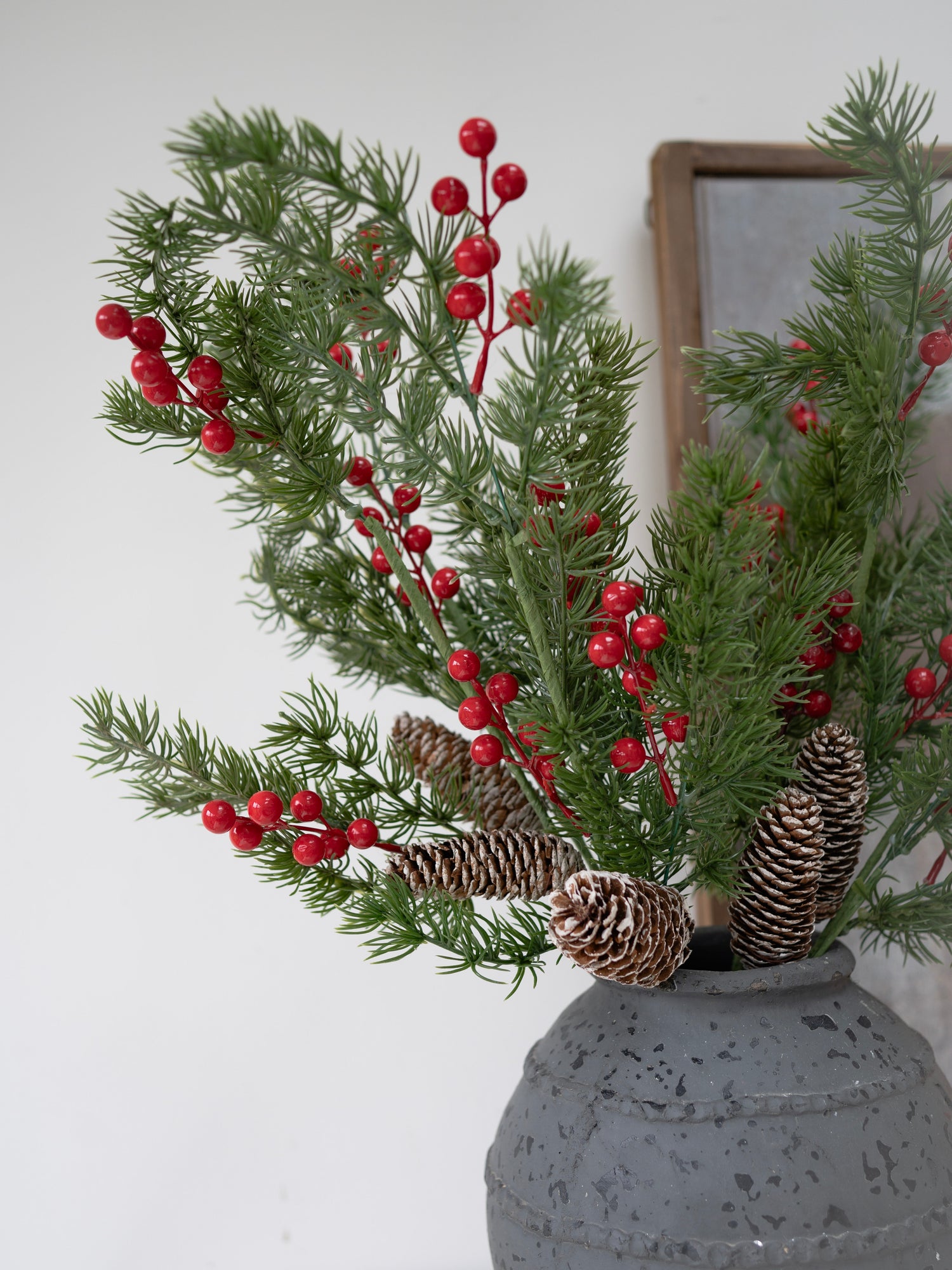 Decorative arrangement with faux christmas greenery, red berries, and pinecones in a gray vase against a white wall.