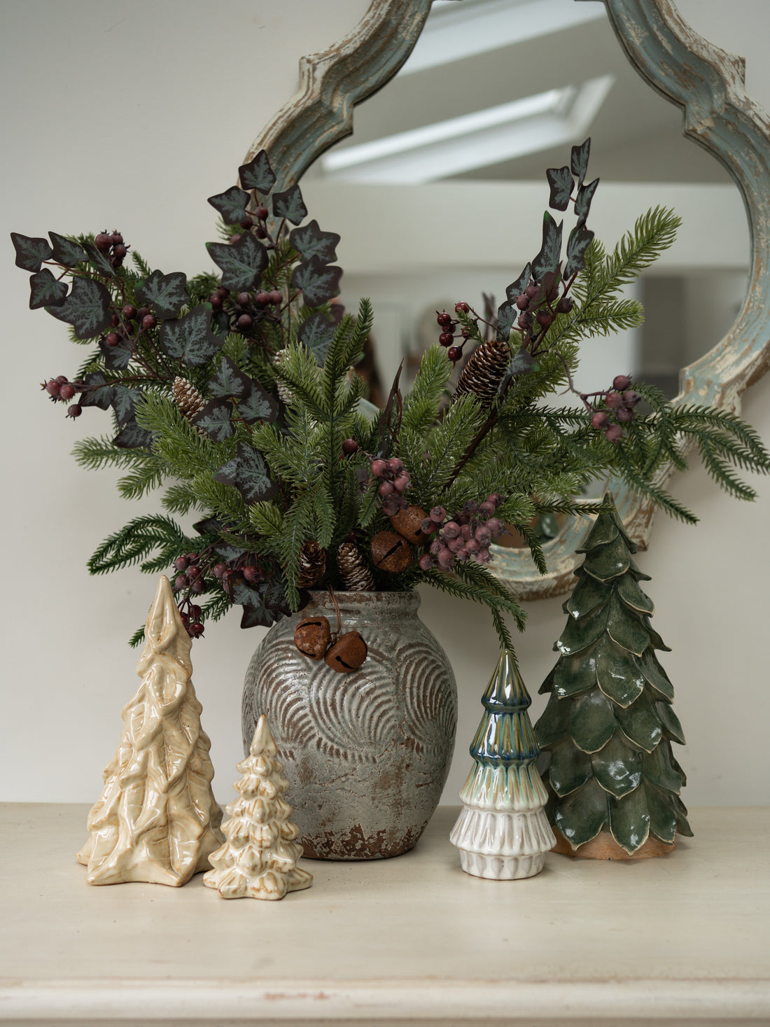 A festive christmas greenery arrangement in a textured gray ceramic vase, styled on a mantelpiece with a distressed mirror, two cream ceramic Christmas trees, and a green ceramic Christmas tree.
