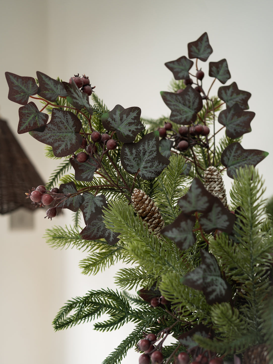 A close-up of a decorative evergreen arrangement featuring dark burgundy ivy with silver undersides, pinecones, and green pine needles against a soft beige background.