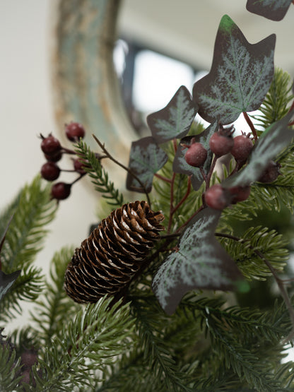 A close-up of a christmas floral arrangement featuring green pine needles, a brown pinecone, dark ivy leaves with silvery-white veining, and clusters of small dusty rose berries.