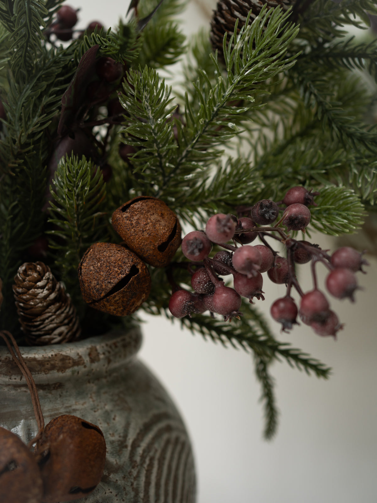 A close-up of a rustic christmas floral arrangement in a textured gray ceramic pot, featuring green pine branches, brown glittery bells, pinecones, and clusters of dusty pink berries against a soft off-white background.