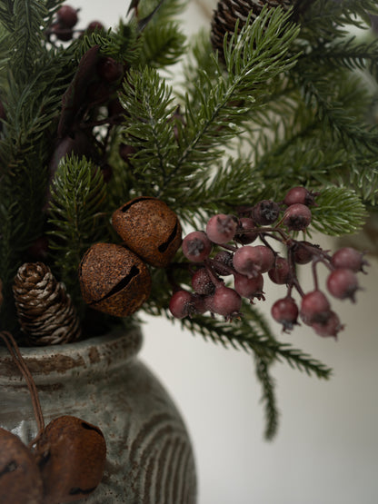 A close-up of a rustic christmas floral arrangement in a textured gray ceramic pot, featuring green pine branches, brown glittery bells, pinecones, and clusters of dusty pink berries against a soft off-white background.