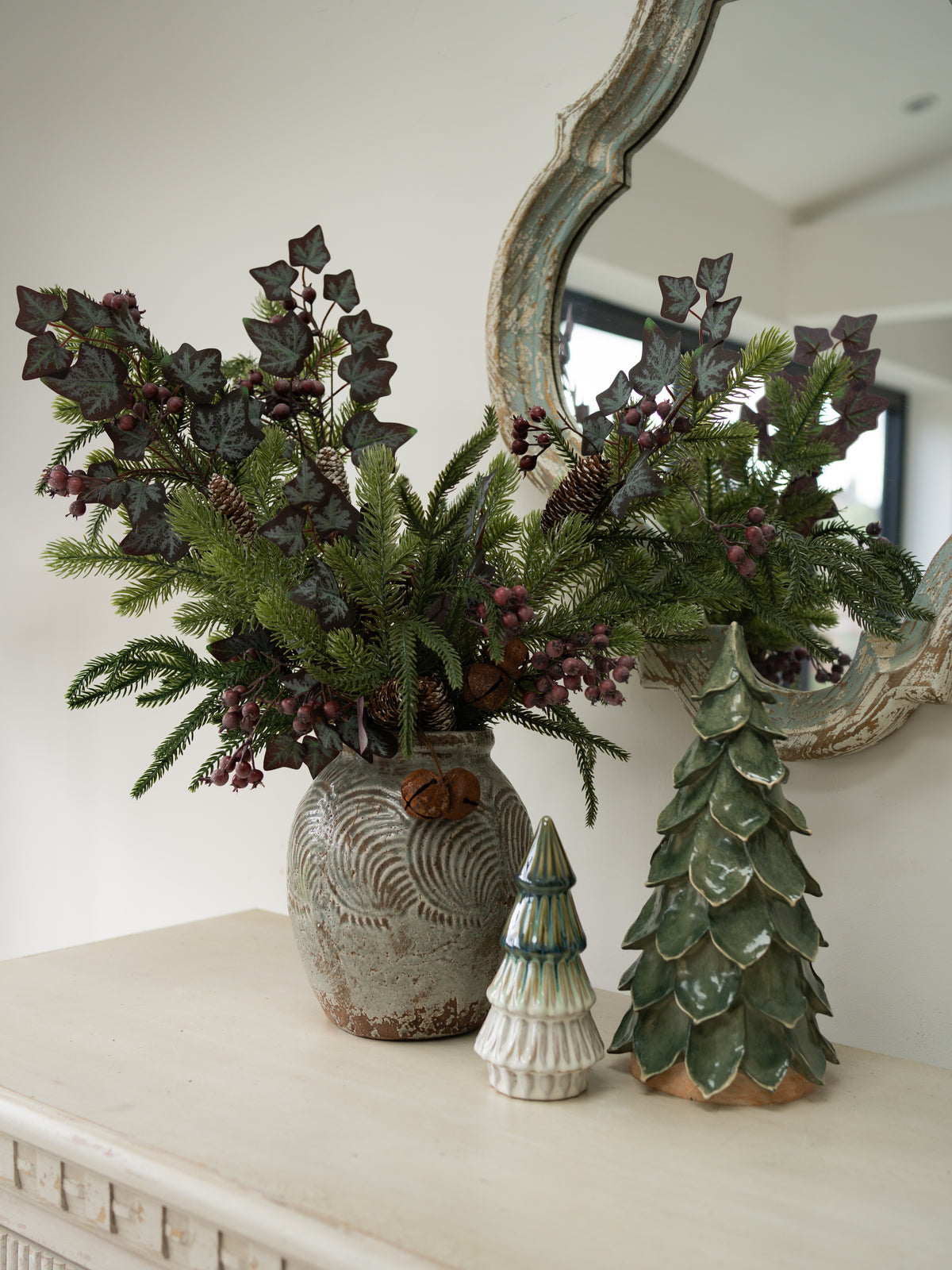 A christmas floral arrangement in a textured gray ceramic vase, featuring green pine branches, dark purple holly leaves, and small berries, styled on a cream mantel next to a green ceramic Christmas tree and a small white figurine.