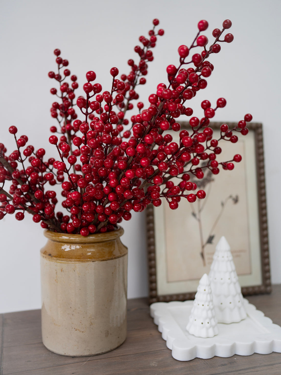 Vase with faux red berries on a wooden surface with a white decorative tree and framed picture in the background.