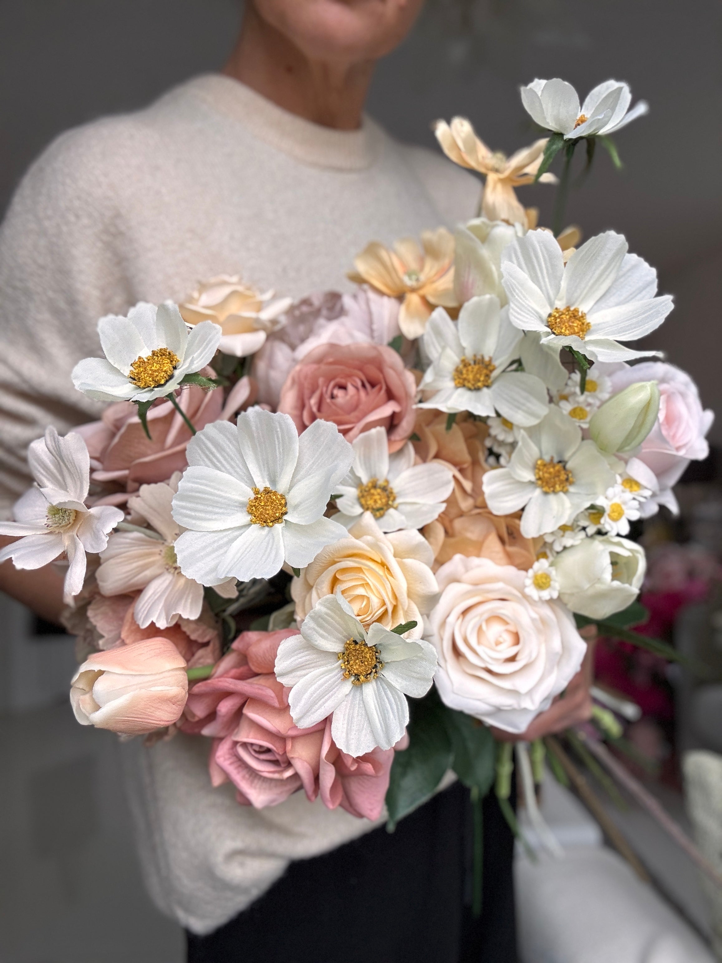 A person in a cream sweater holds a large bouquet of artificial flowers, including white daisy-like blooms with yellow centers, blush pink roses, and pale peach roses.