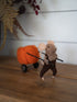 A felted wool mouse figurine dressed in brown overalls and a beige shirt, pushing a small orange pumpkin wagon with black wheels on a wooden surface.
