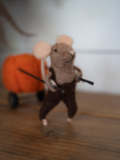 A handcrafted felted wool mouse figurine wearing brown pants and holding a black wand, standing on a wooden surface with a blurred orange pumpkin cart in the background.