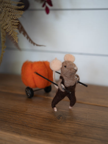 A handcrafted felted wool mouse figurine wearing brown overalls and pushing an orange felted pumpkin wagon with black wheels, set on a wooden surface against a white paneled wall.