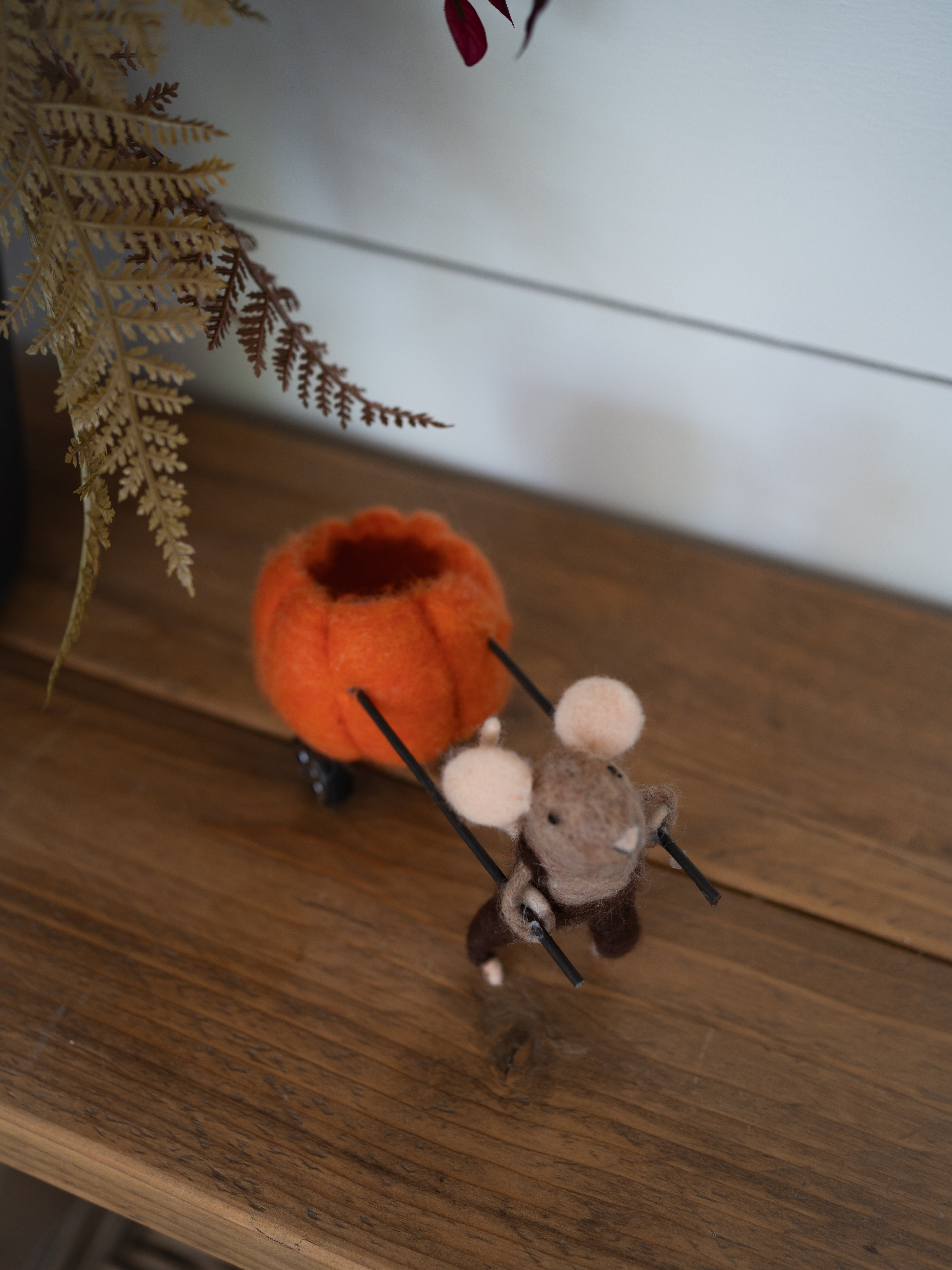 A handcrafted felted wool mouse pulling a small orange pumpkin on a wooden surface, with dried fern leaves in the background.