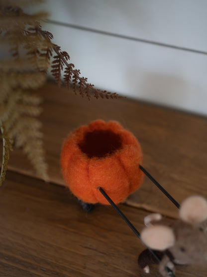 A small orange felt pumpkin-shaped container or candle holder with a dark opening, resting on a wooden surface with dried fern leaves and a small plush mouse nearby.