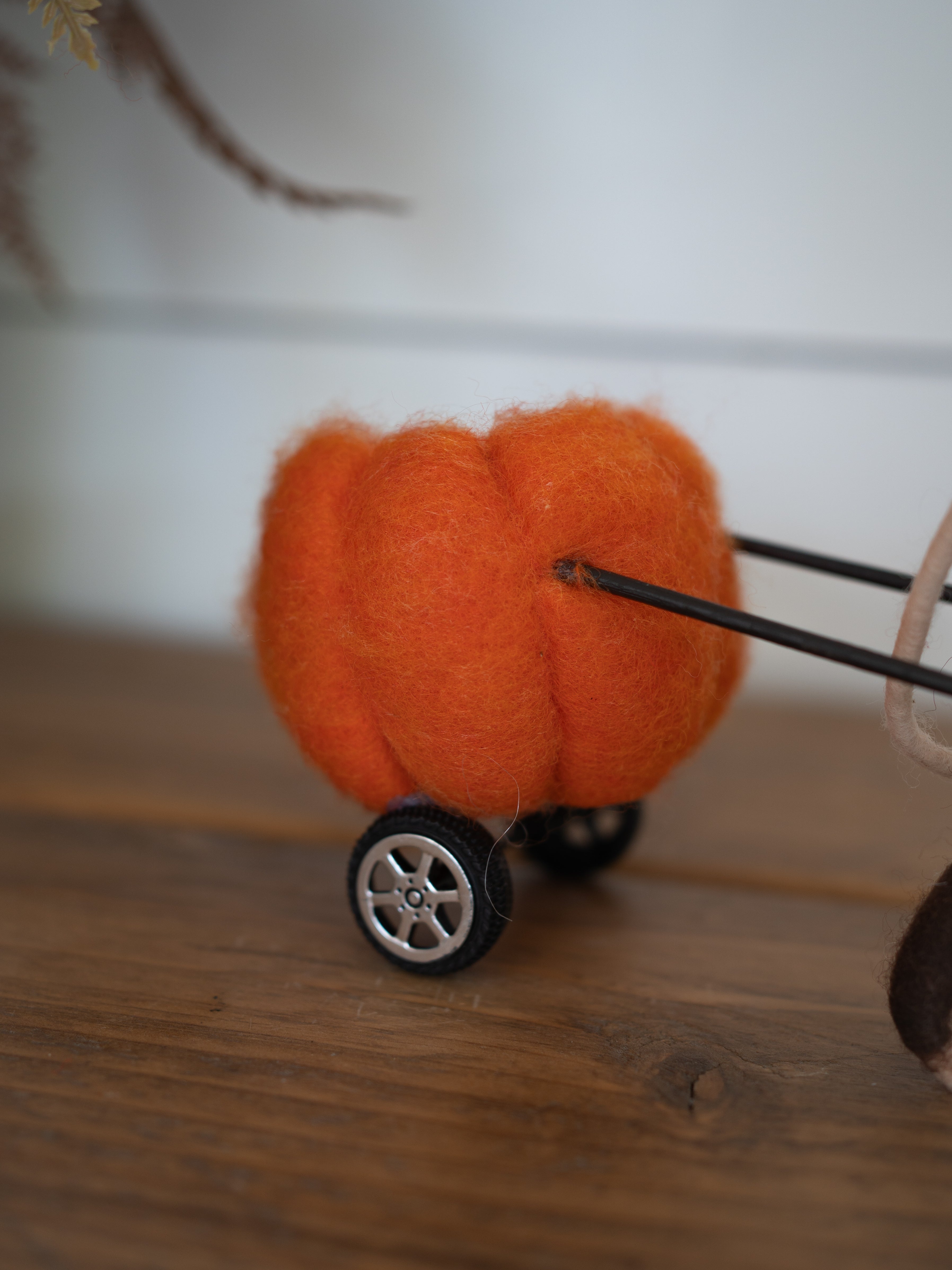 A small orange felt pumpkin decoration on a miniature black and silver toy cart, resting on a wooden surface with a soft-focus indoor background.
