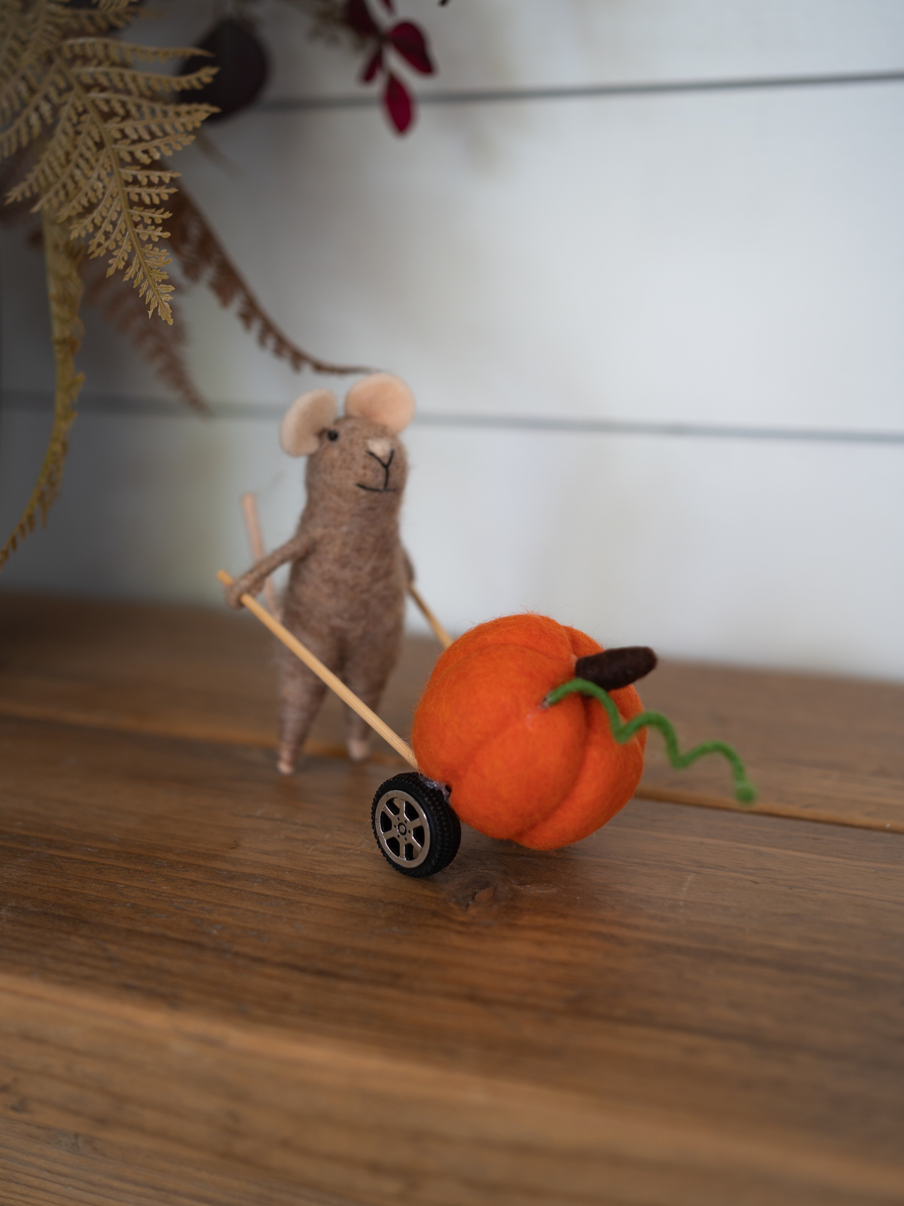 A small felted wool mouse figurine pushes a miniature orange pumpkin cart with a green stem and black wheel, set on a wooden surface against a white paneled background.