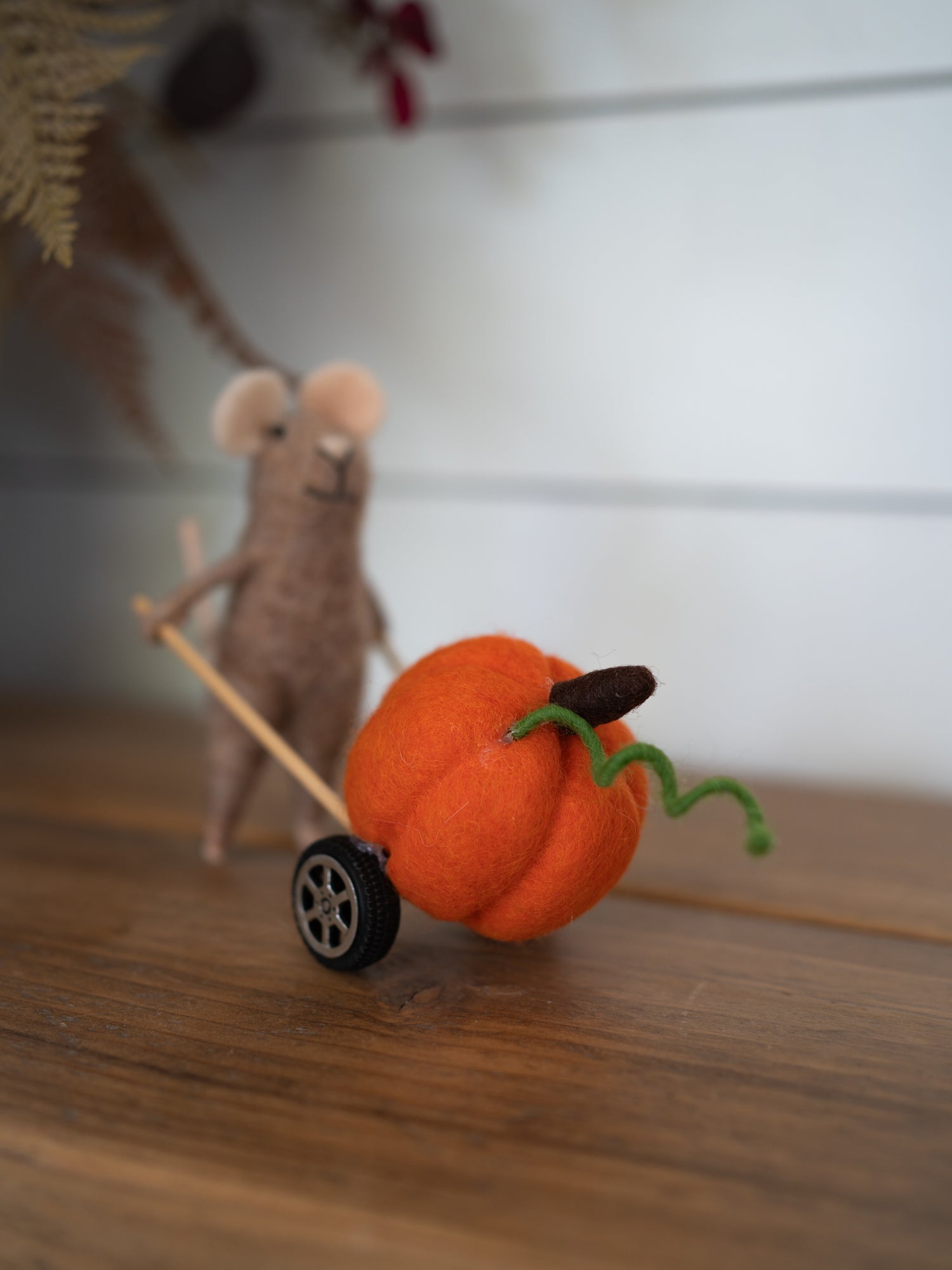 A small orange felt pumpkin decoration with a green curly stem and a dark brown stem, pushed by a beige felt mouse on a wooden surface.