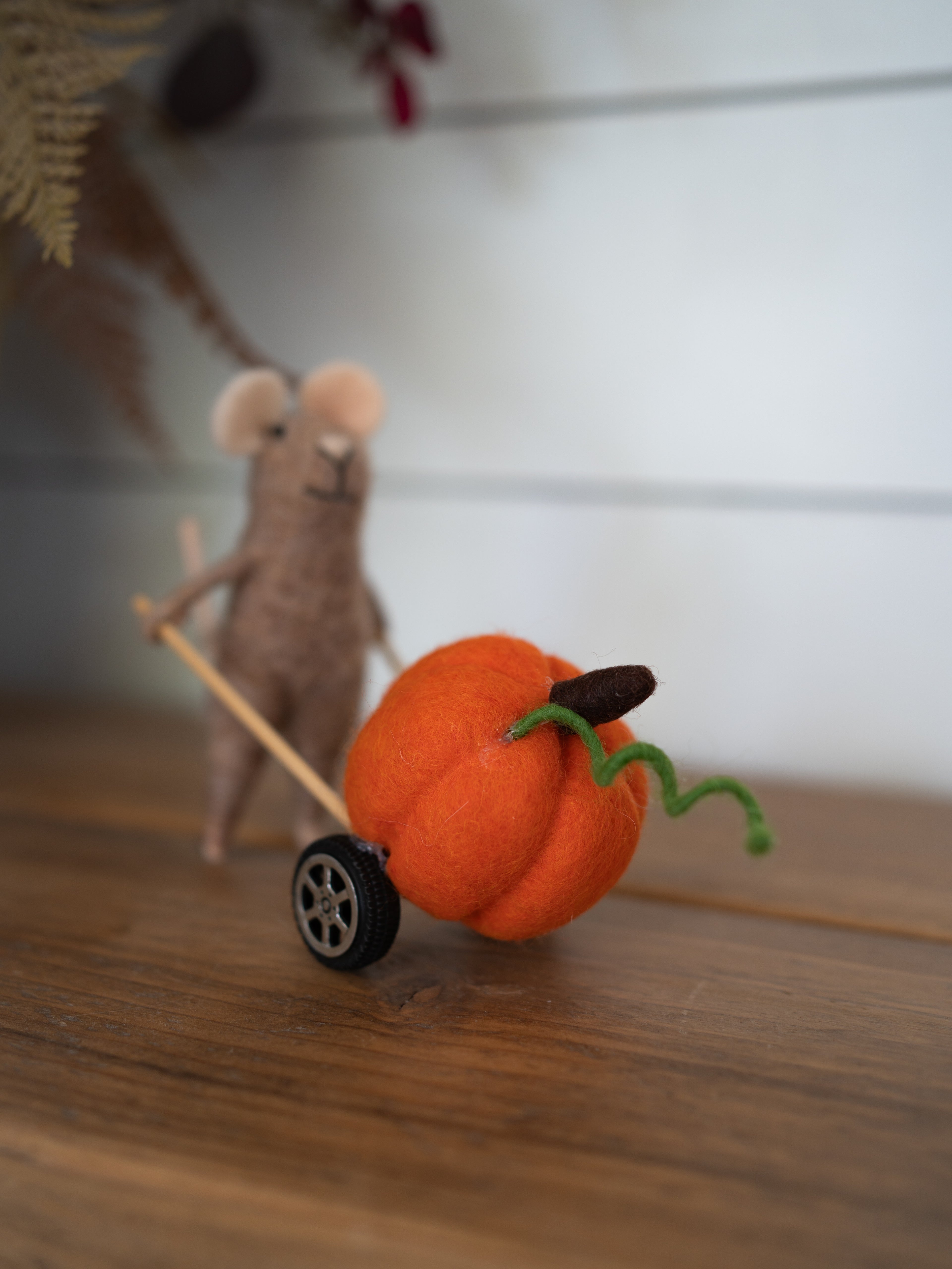 A small orange felt pumpkin decoration with a green curly stem and a dark brown stem, pushed by a beige felt mouse on a wooden surface.