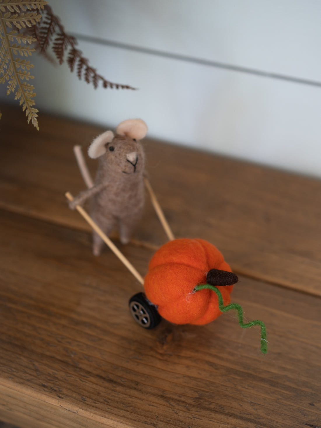 A handcrafted felted wool decoration featuring a small gray mouse pushing a miniature orange pumpkin cart with a green wire stem, set on a wooden tabletop.