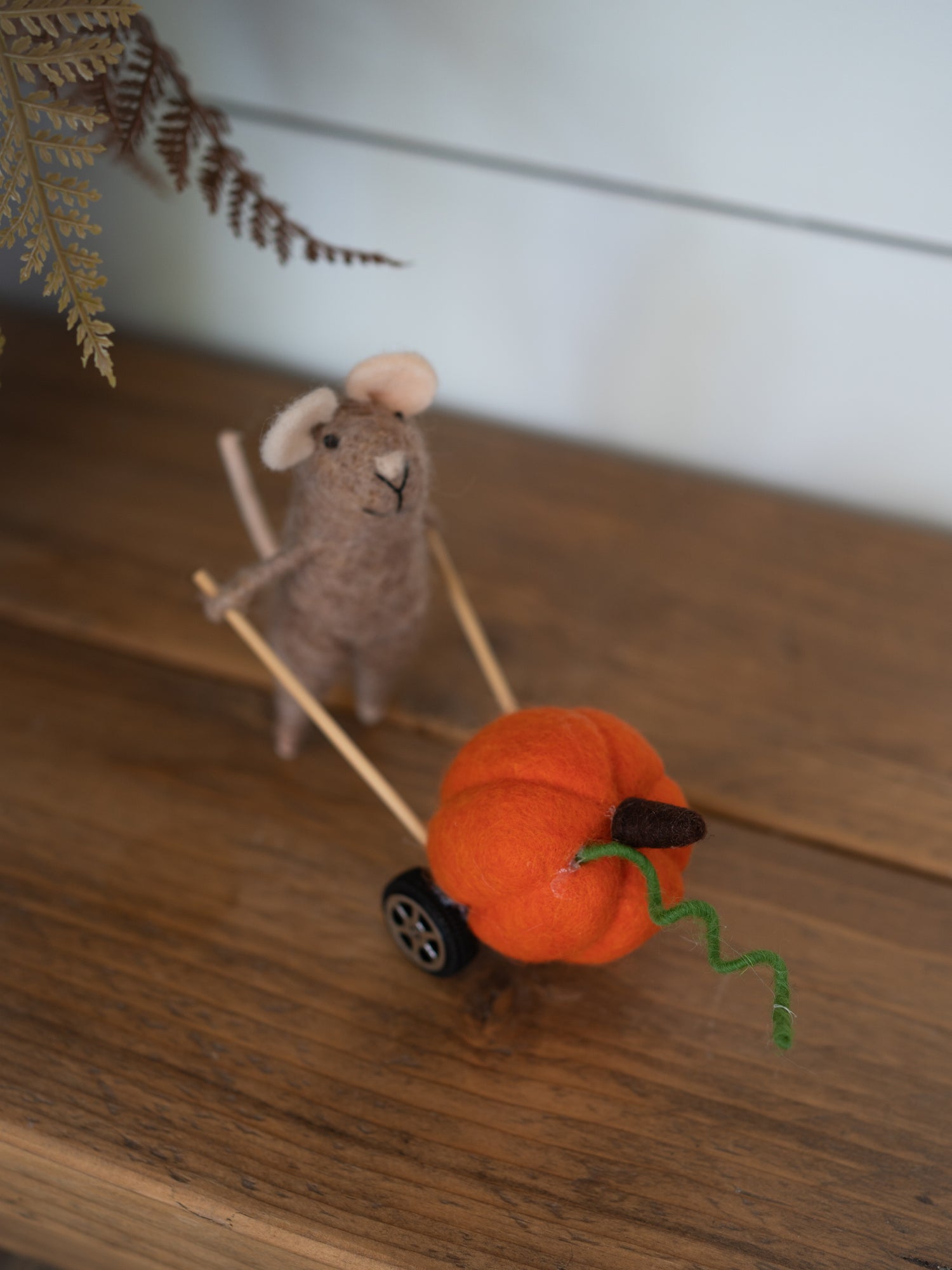 A handcrafted felted wool decoration featuring a small gray mouse pushing a miniature orange pumpkin cart with a green wire stem, set on a wooden tabletop.