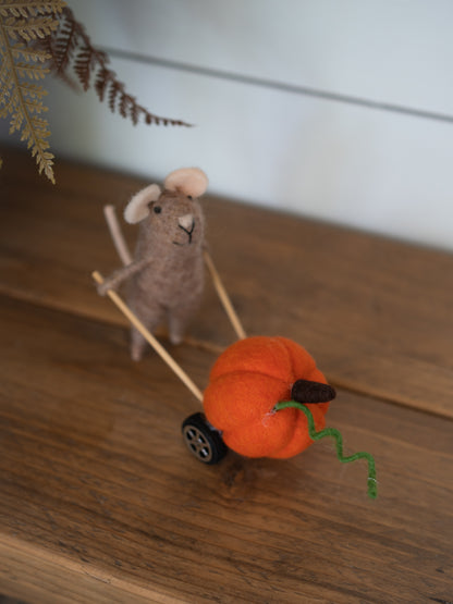 A handcrafted felted wool decoration featuring a small gray mouse pushing a miniature orange pumpkin cart with a green wire stem, set on a wooden tabletop.