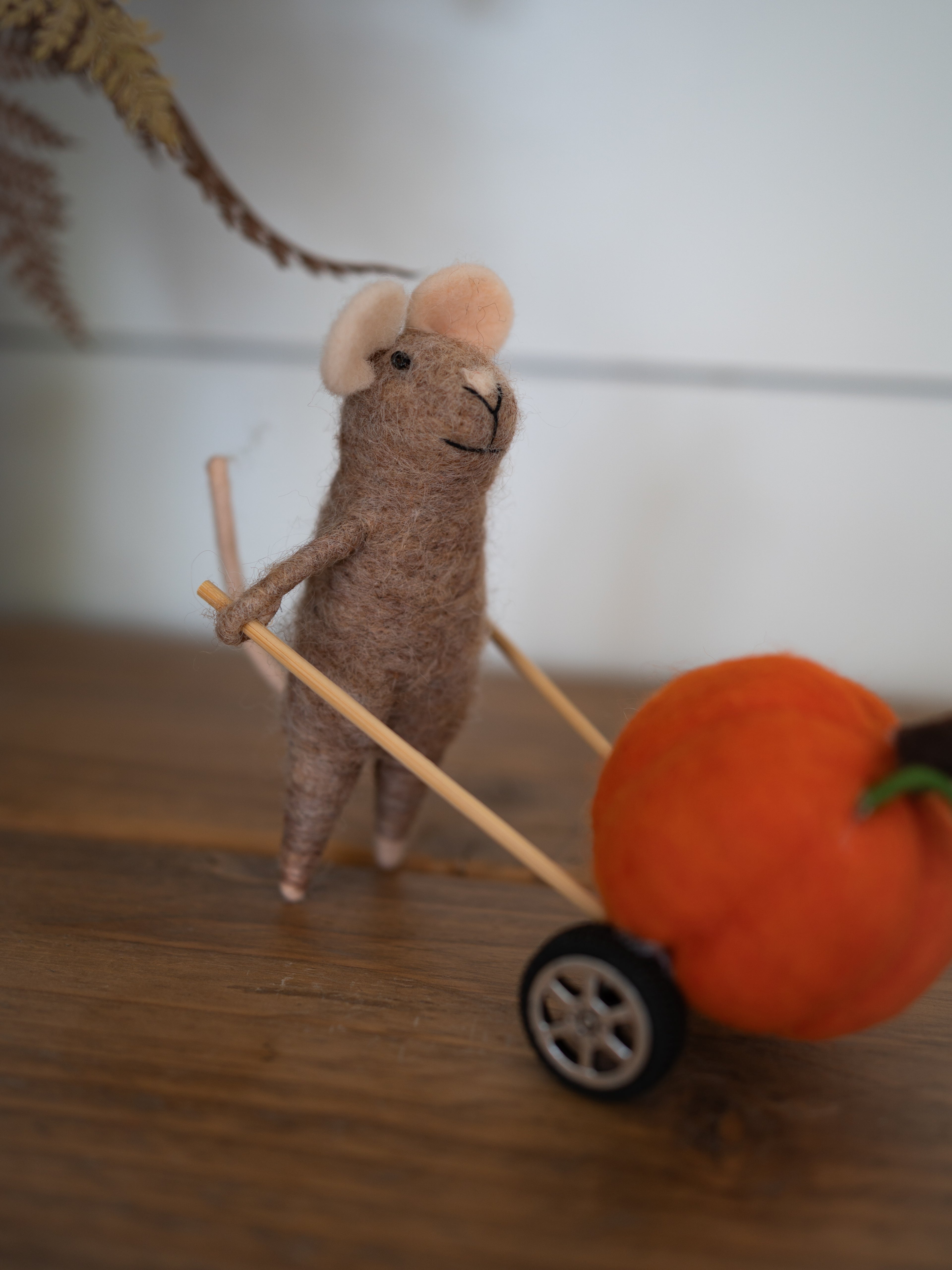 A felted wool mouse toy pushing a small orange pumpkin with a black wheel, set on a wooden surface against a soft-focus indoor background.