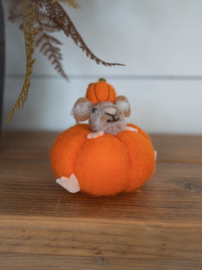 A small felted wool mouse figurine sitting on top of an orange pumpkin, with a smaller orange pumpkin on its head, displayed on a wooden surface.