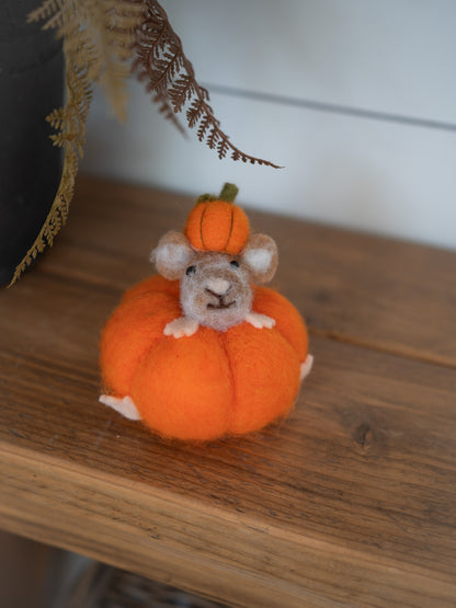 A small felted wool mouse figurine sitting inside an orange pumpkin, displayed on a wooden shelf.