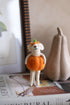 Small plush toy dressed in a pumpkin costume on a surface with a newspaper and gourd in the background.