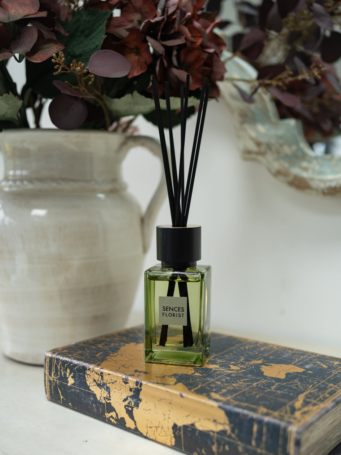 A green glass sences florist reed diffuser bottle with black reeds and a black cap sits on a gold-and-blue patterned book, with a cream ceramic vase and dried burgundy foliage blurred in the background.