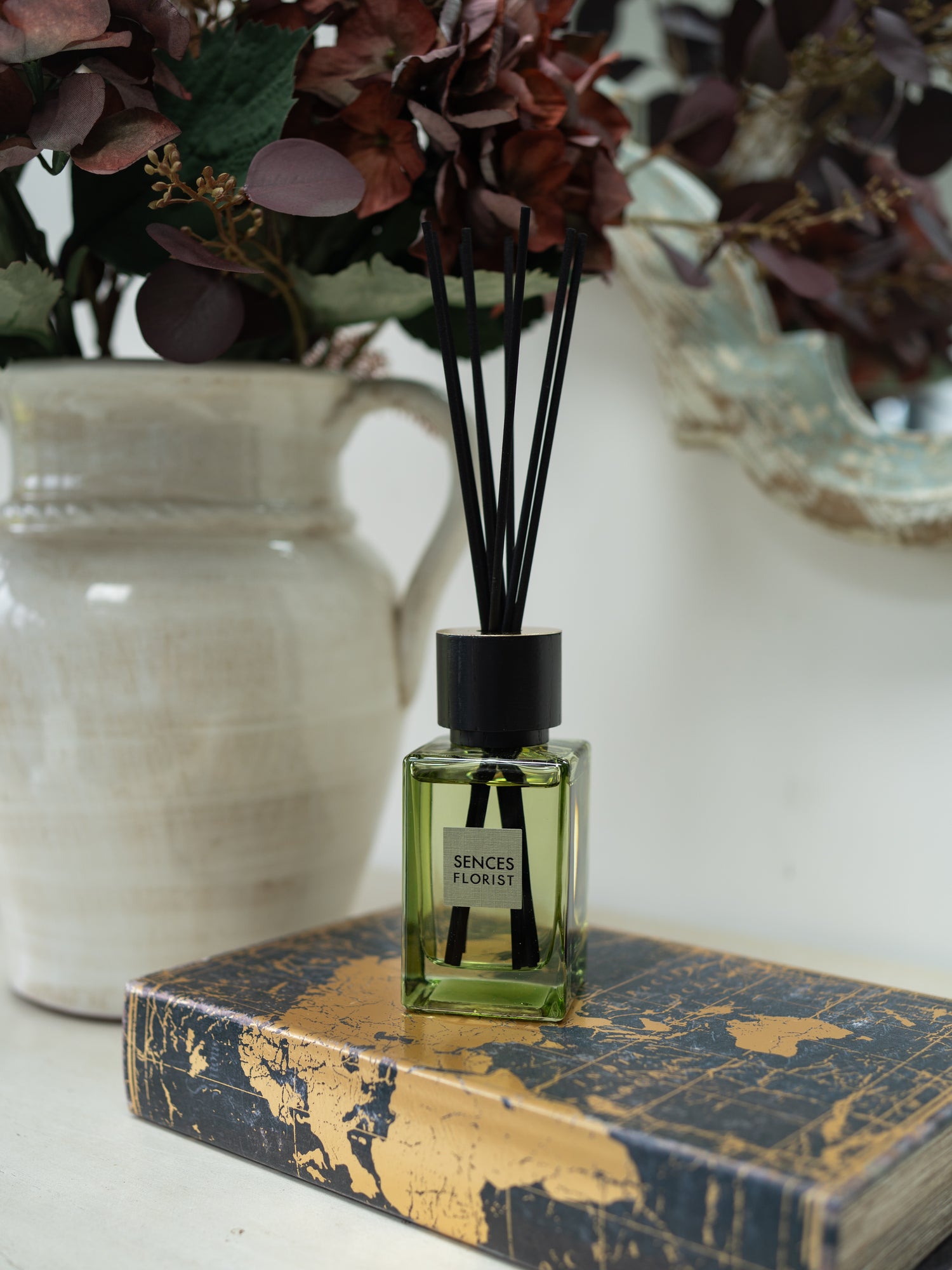 A green glass sences florist reed diffuser bottle with black reeds and a black cap sits on a gold-and-blue patterned book, with a cream ceramic vase and dried burgundy foliage blurred in the background.